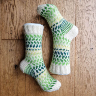 A pair of green, yellow and cream colourwork socks on foot forms lying on a wooden floor. The feet are positioned so that the sock toes are opposite each other