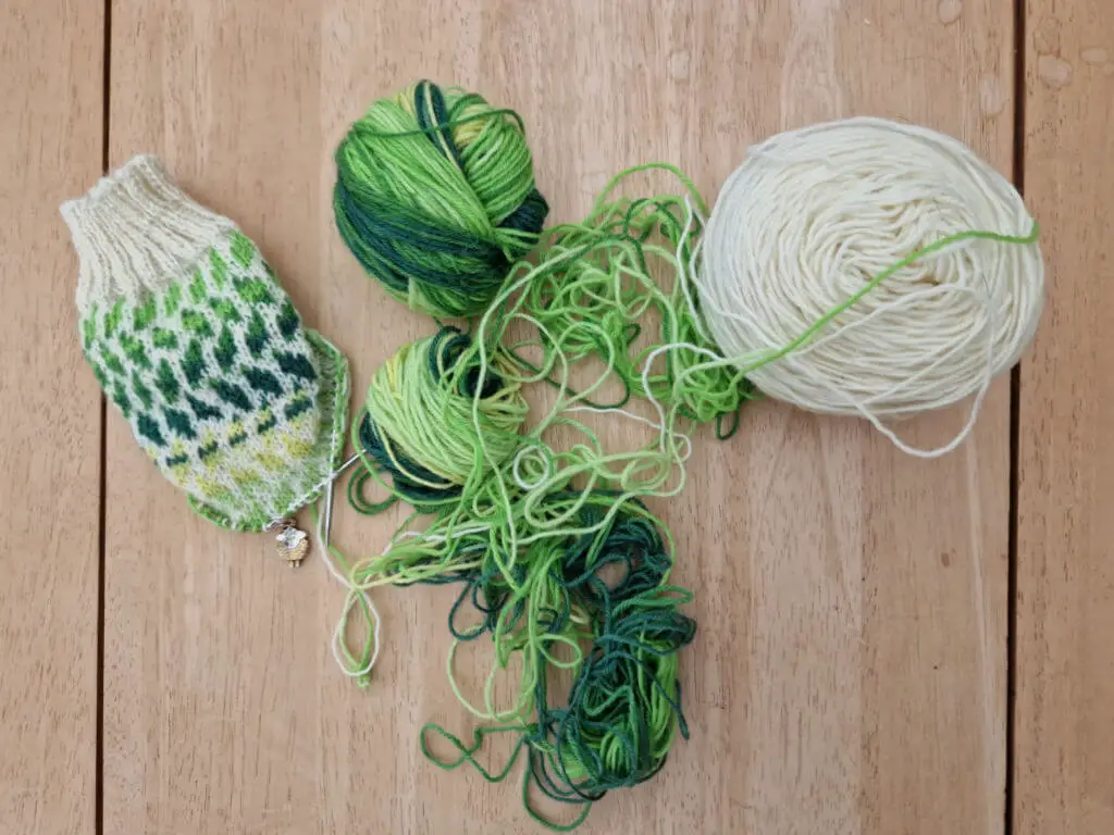 A partly-knitted colourwork sock in cream and green-striped yarn. There is a mess of green yarn on the table