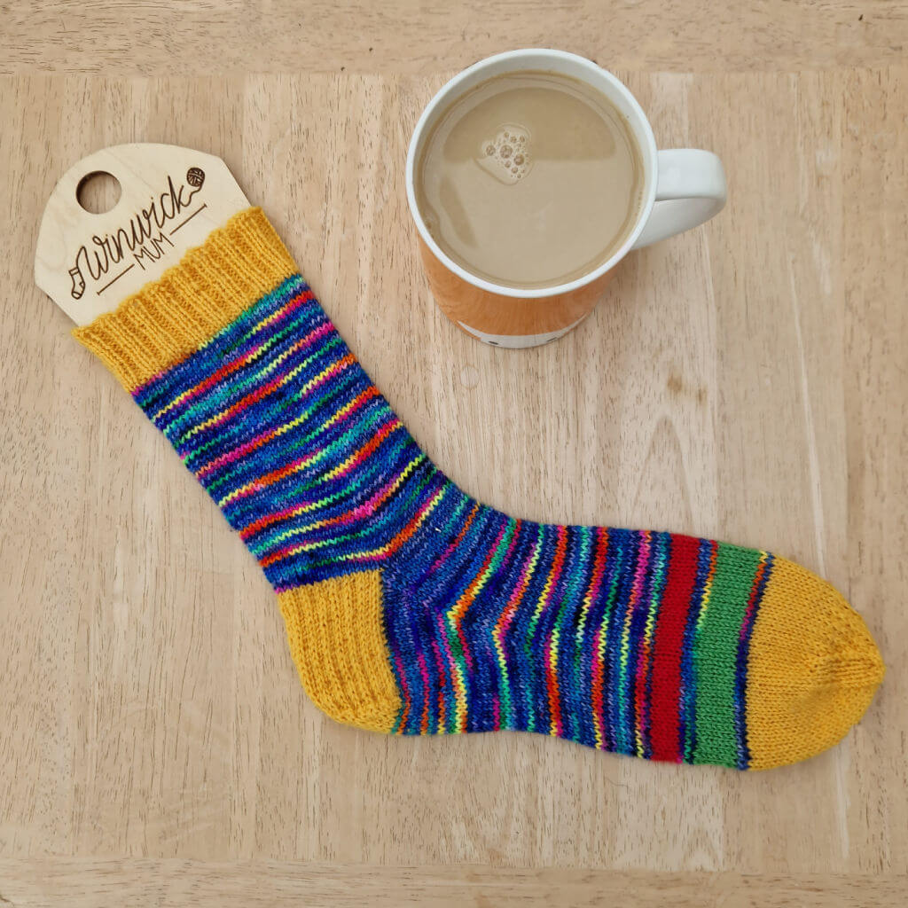A hand knitted sock in rainbow yarn with stripes of red, green and yellow at the bottom of the foot. The cuff and heel is yellow. The sock is on a wooden sock blocker, lying on a wooden table next to an orange mug of tea