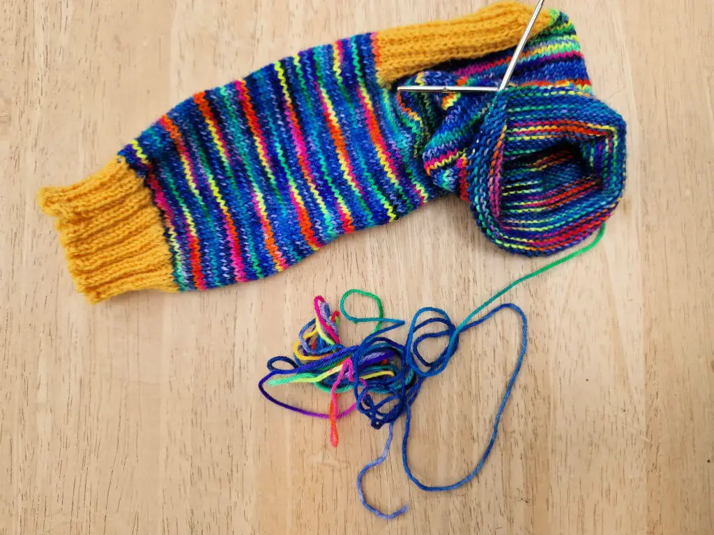 A partly-knitted sock in rainbow colours with a yellow cuff and heel flap