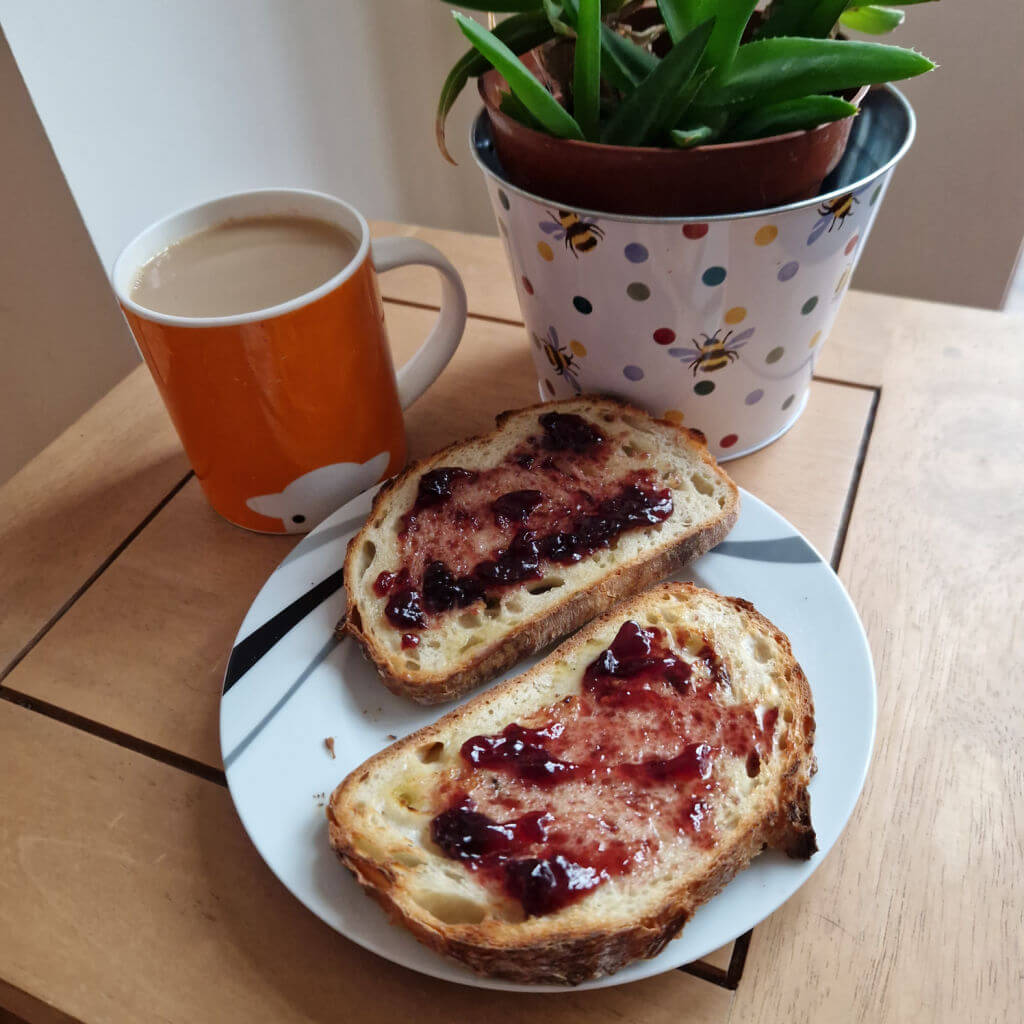 A wooden coffee table with a plant in a spotty plant pot, an orange mug of tea and two slices of toast and jam on a plate