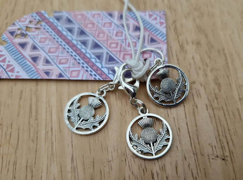 Three stitch markers in the shape of a thistle within a circle attached to a piece of coloured card