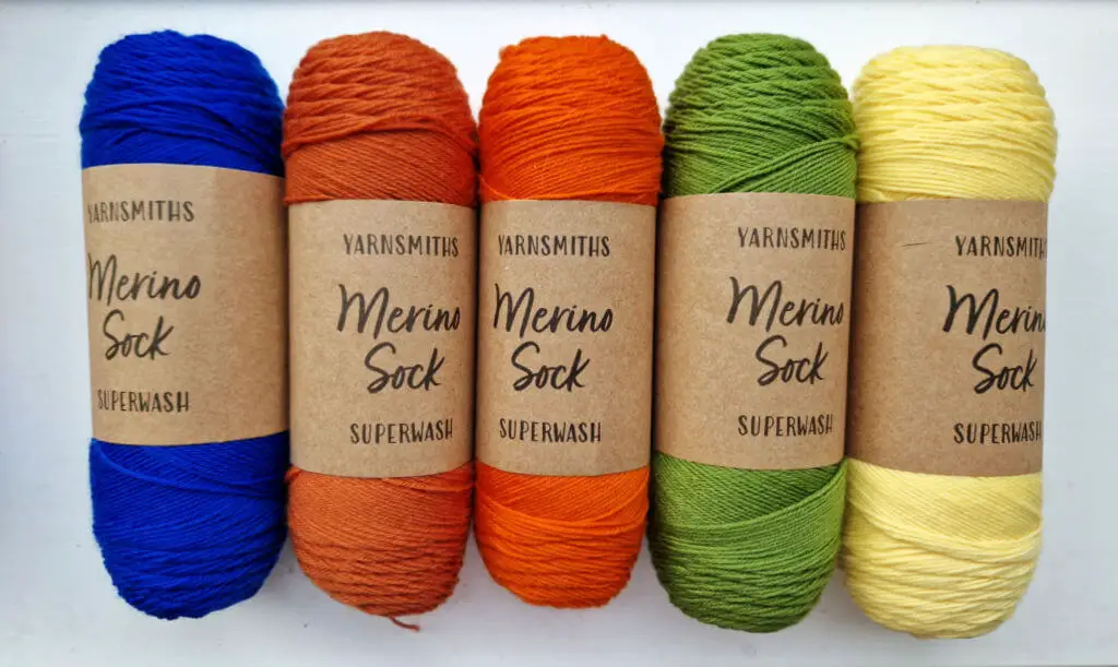 Five balls of sock yarn in blue, cinnamon, pumpkin, green and yellow colours lined up on a white background