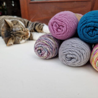 Five balls of sock yarn piled on top of each other with a tabby cat rolling on the floor behind them