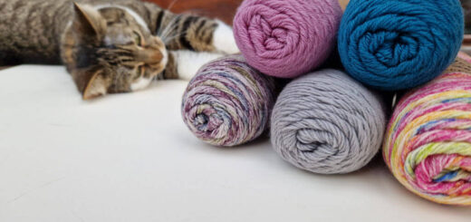 Five balls of sock yarn piled on top of each other with a tabby cat rolling on the floor behind them