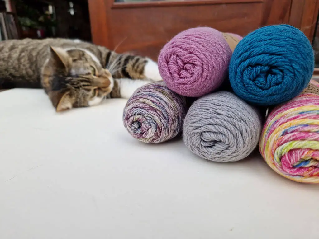 Five balls of sock yarn piled on top of each other with a tabby cat rolling on the floor behind them