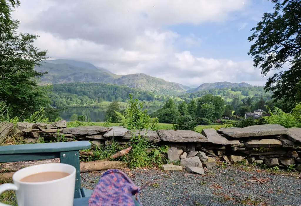 A view across a gravel area bordered by a dry stone wall to the mountains beyond. A partly-knitted sock and a white mug of tea are balanced on the arm of an outdoor chair
