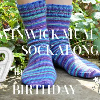 A pair of hand knitted blue striped socks modelled on feet which are standing on a stone flag in a flower border. The text across the picture reads Winwick Mum Sockalong 9th birthday