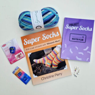 An orange Super Socks beginner sock knitting book, a purple More Super Socks notebook, a ball of yarn in shades of blue, and white, a row counter, a packet of wool needles and three stitch markers on a white background