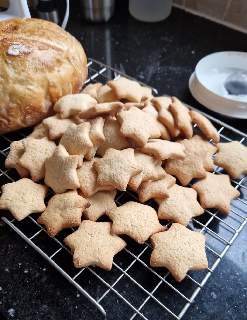 A pile of star-shaped biscuits (cookies) on a wire cooling rack next to a freshly-baked loaf of bread