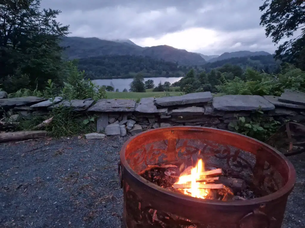 A small fire burns in a fire basket with a view across a gravel area bordered by a dry stone wall to the mountains beyond. 