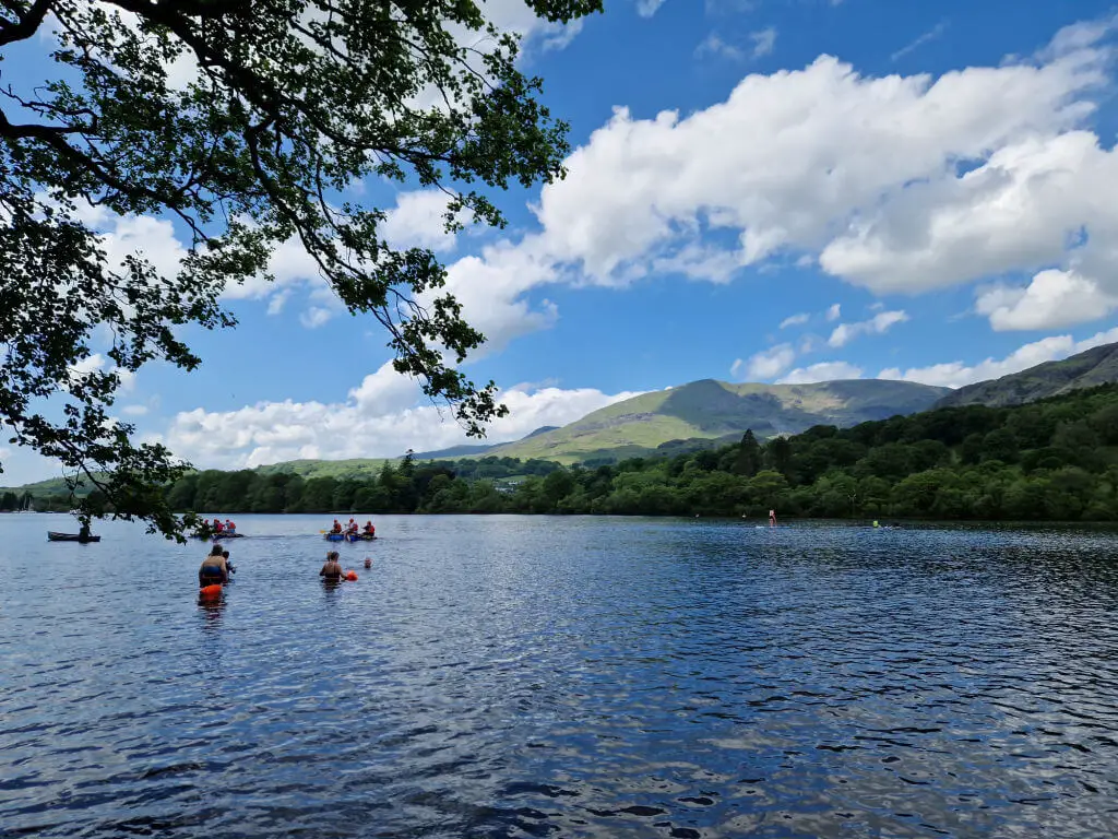 A view across a lake to the hills beyond. There are people swimming in the lake. The sky is blue and bright