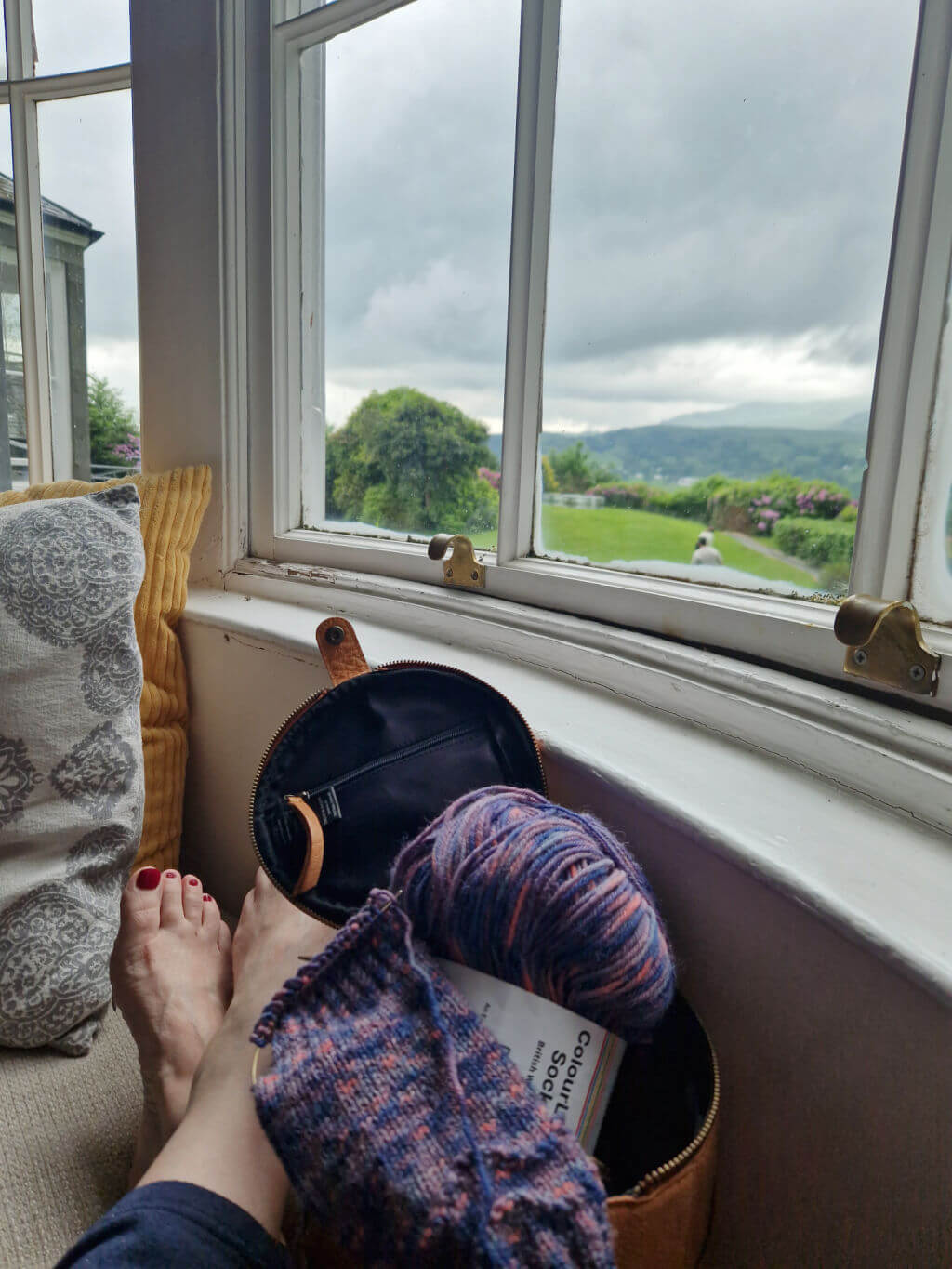 Christine is sitting on a window seat with her feet up on the cushions. Her feet are bare and crossed at the ankle. There is a round leather project bag and partly knitted sock on the seat next to her, and through the window you can see across the lawn to the hills beyond.