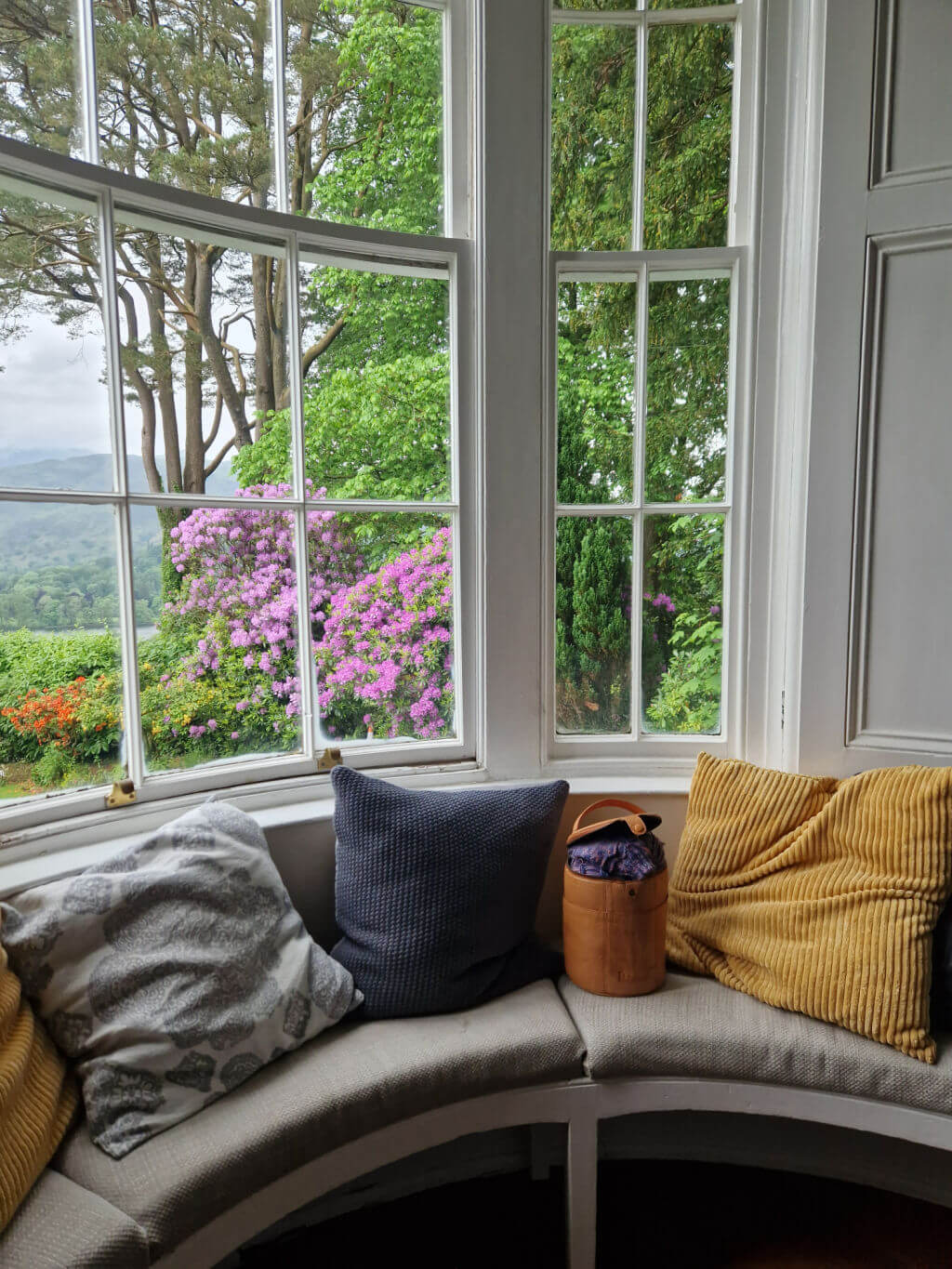 A window seat in a round bay window. There are square cushions on the seat and a round leather sock project bag
