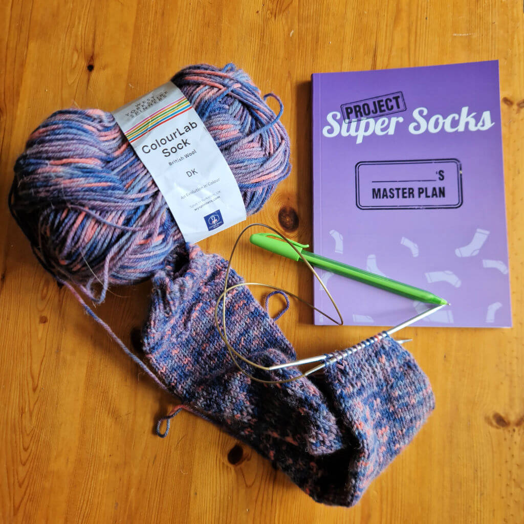 A nearly-finished hand knitted sock next to a ball of yarn and a purple sock notebook on a wooden table