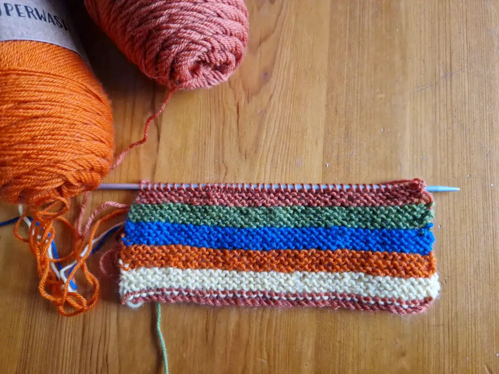 A piece of knitting on a needle in stripes of yellow, green, blue, brown and pumpkin colours