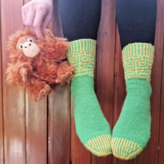 A pair of green and yellow patterned socks modelled on feet. The wearer is sitting with her legs over the edge of a wooden wall so that she is suspended above the ground. There's a monkey face on the socks, and the wearer is holding a small toy monkey