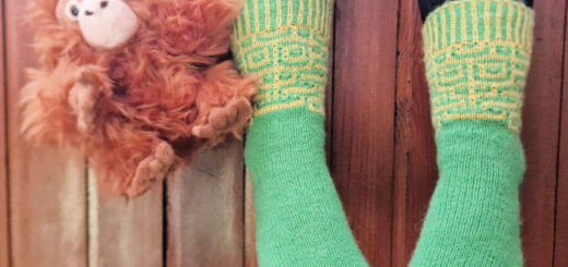 A pair of green and yellow patterned socks modelled on feet. The wearer is sitting with her legs over the edge of a wooden wall so that she is suspended above the ground. There's a monkey face on the socks, and the wearer is holding a small toy monkey