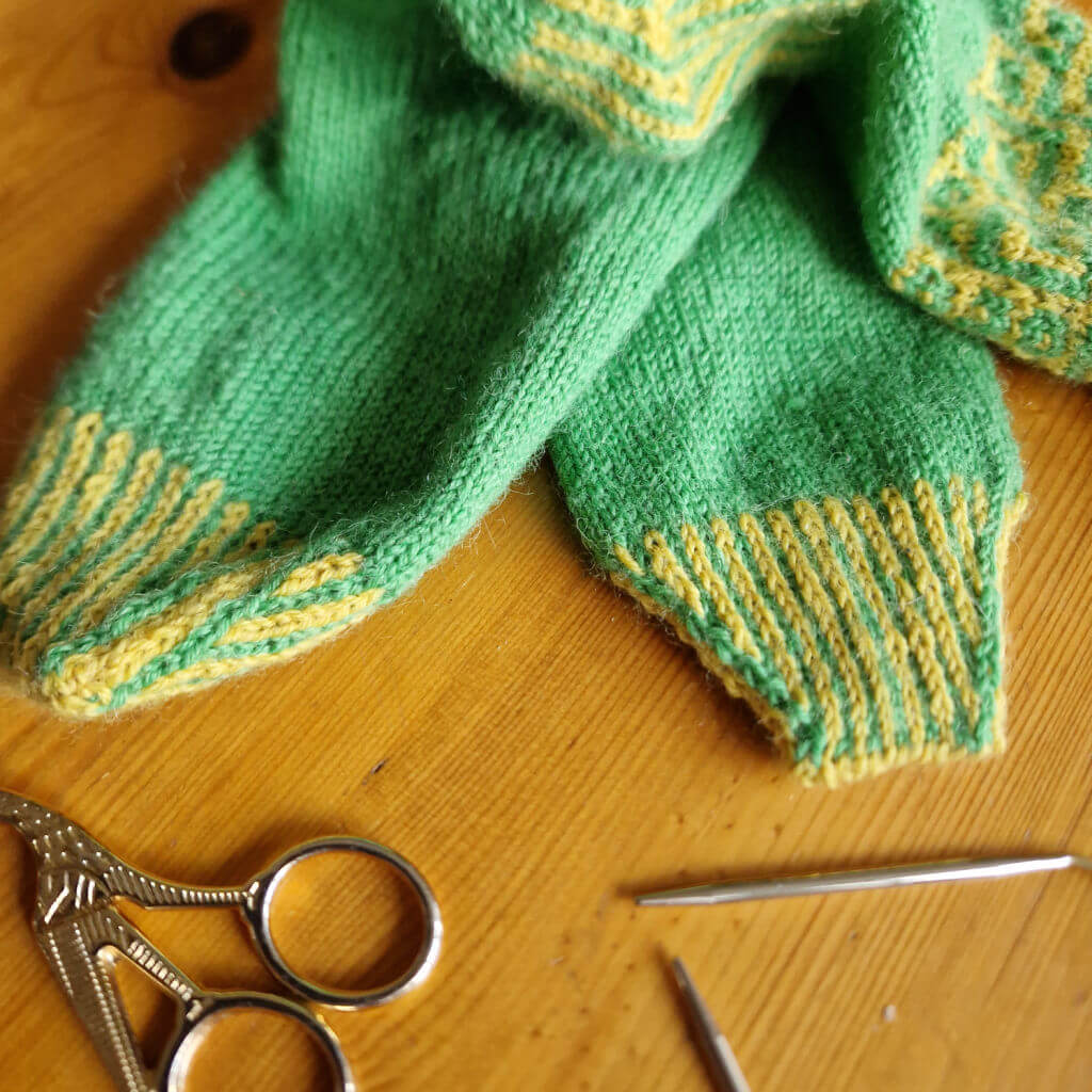 A pair of green and yellow socks on a wooden table next to a small pair of gold scissors and a short circular knitting needle