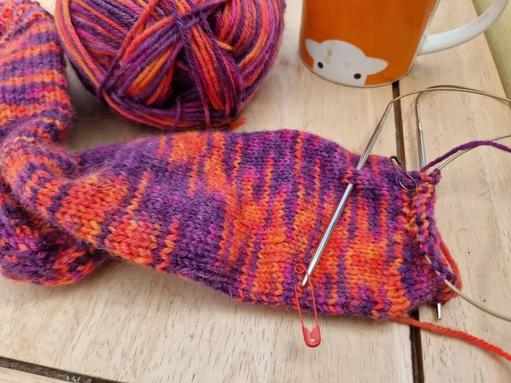 A partly-knitted sock in shades of pink, red and purple on a wooden coffee table next to an orange mug of tea