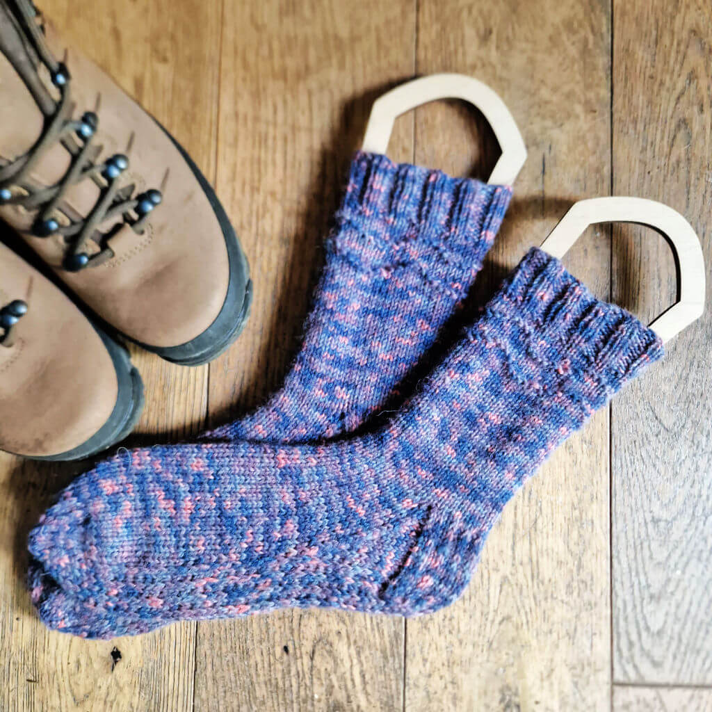 A pair blue variegated socks on sock blockers lying on a wooden floor next to a pair of hiking boots
