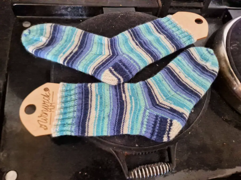 A pair of blue, turquoise and white striped socks on wooden sock blockers drying on the round lid of an Aga hot plate