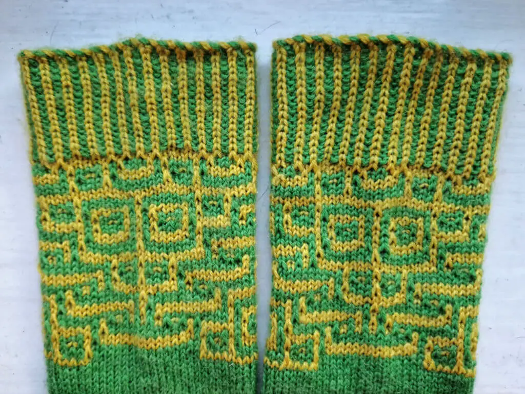 Two green and yellow socks with a mosaic pattern featuring a monkey face lie side by side on a white background