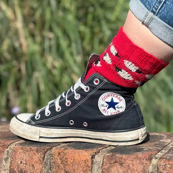 A red colourwork sock featuring black and white sheep, modelled on a foot, is worn with a black and white canvas boot