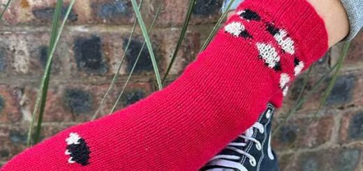 A red colourwork sock featuring black and white sheep, modelled on a foot, is resting over a black and white canvas boot