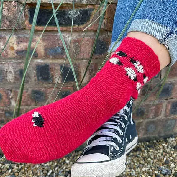 A red colourwork sock featuring black and white sheep, modelled on a foot, is resting over a black and white canvas boot