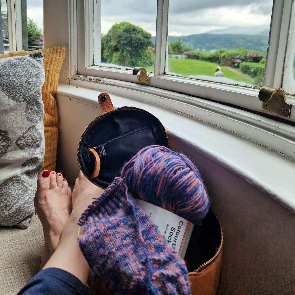 Christine is sitting on a window seat with her feet up on the cushions. Her feet are bare and crossed at the ankle. There is a round leather project bag and partly knitted sock on the seat next to her, and through the window you can see across the lawn to the hills beyond.