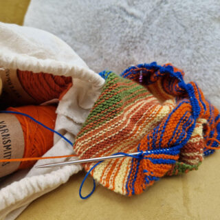 A multi-coloured partly-knitted shawl on a mustard coloured sofa next to a fabric bag of yarn