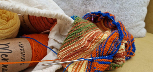 A multi-coloured partly-knitted shawl on a mustard coloured sofa next to a fabric bag of yarn