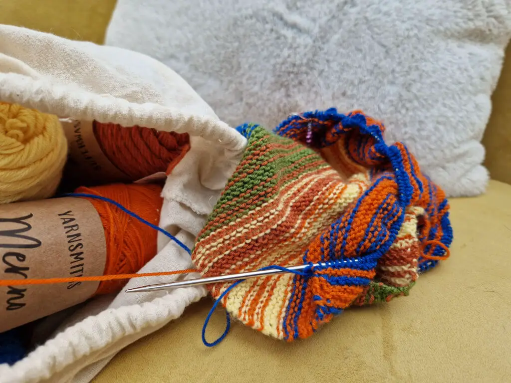 A multi-coloured partly-knitted shawl on a mustard coloured sofa next to a fabric bag of yarn