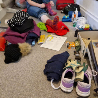 A young woman is sitting on a carpeted floor surrounded by clothes and equipment for a trekking expedition. She is putting a pair of pink hand knitted walking socks on her feet