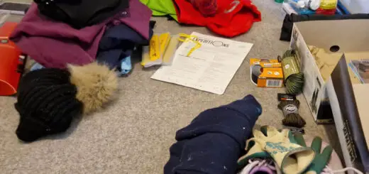 A young woman is sitting on a carpeted floor surrounded by clothes and equipment for a trekking expedition. She is putting a pair of pink hand knitted walking socks on her feet