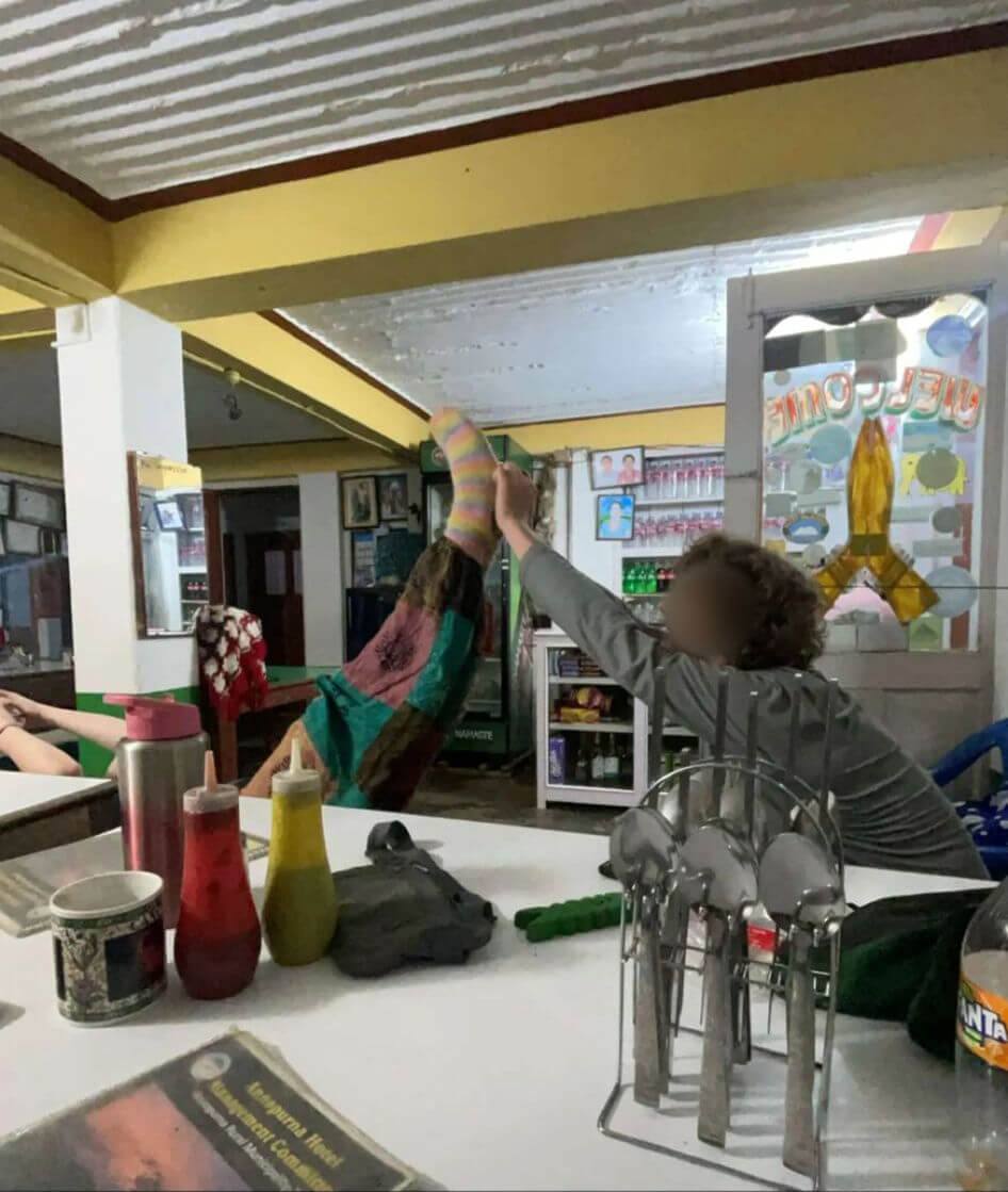 Not so small daughter is lying on a bench next to a dining table with her leg in the air, supported by a friend. She is wearing brightly coloured pants and a rainbow striped hand-knitted sock