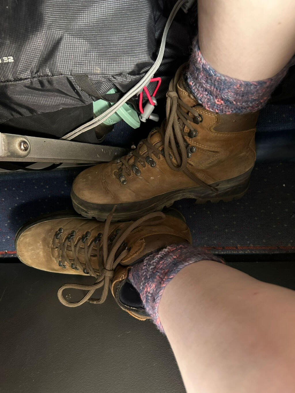 A pair of hand knitted socks in walking boots. The photo is taken from a cramped seat on a plane