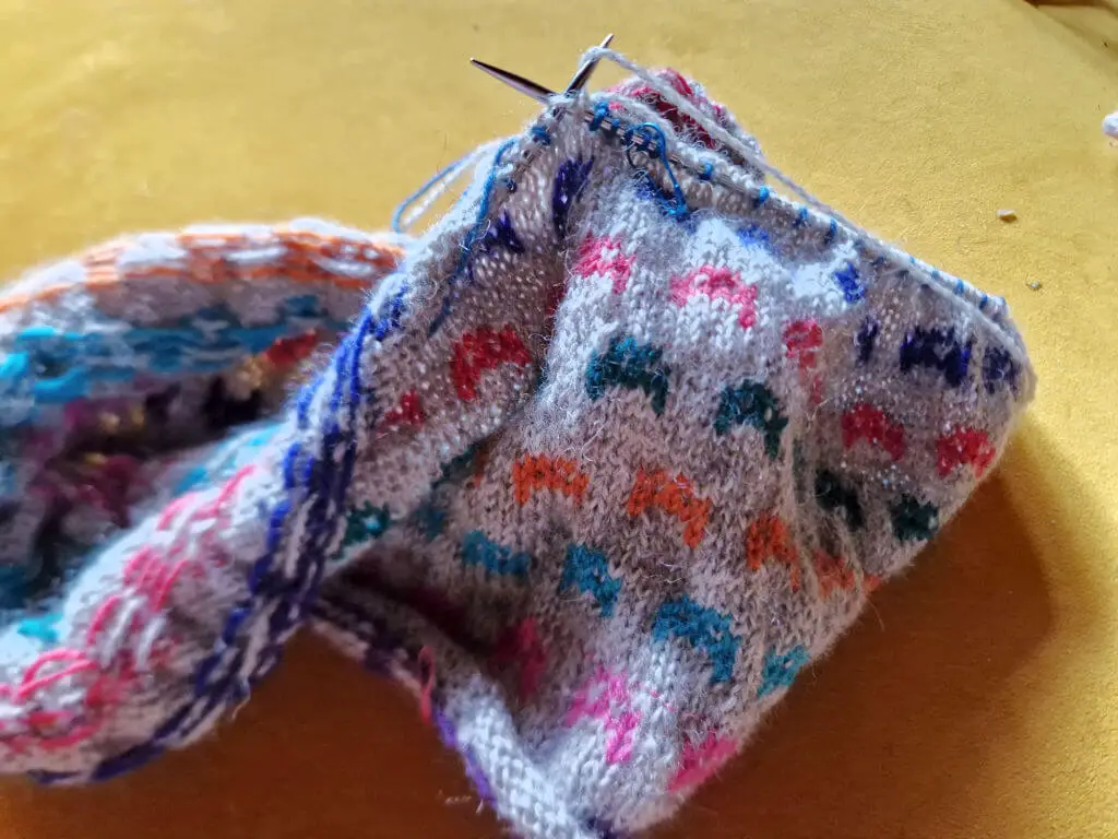 A partly knitted jumper with colourwork cat faces lying on a yellow sofa cushion