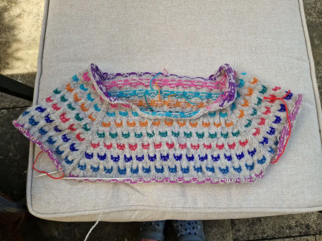 A partly-knitted jumper with colourwork cat faces spread out on a garden chair cushion to show the lines of cats and the jumper shape