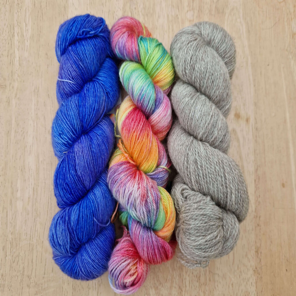 Three skeins of yarn lying on a wooden table. The left hand one is blue, the middle is rainbow and the right hand one is grey