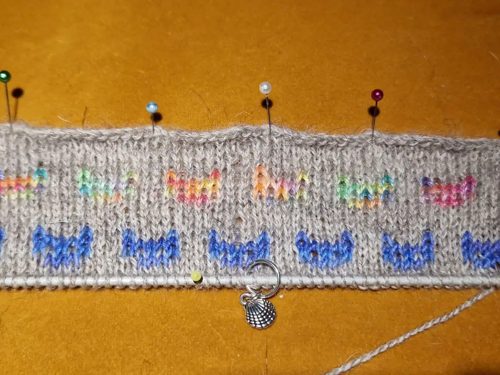 A narrow band of knitting pinned to a mustard coloured cushion cover. The background yarn is grey and there are two rows of knitted cat faces, one rainbow and one blue