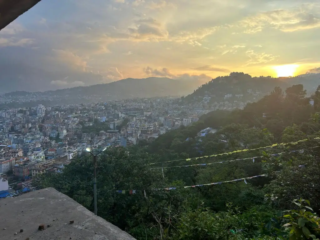 A view across a city from a high point; in the background are hills and the sun is setting
