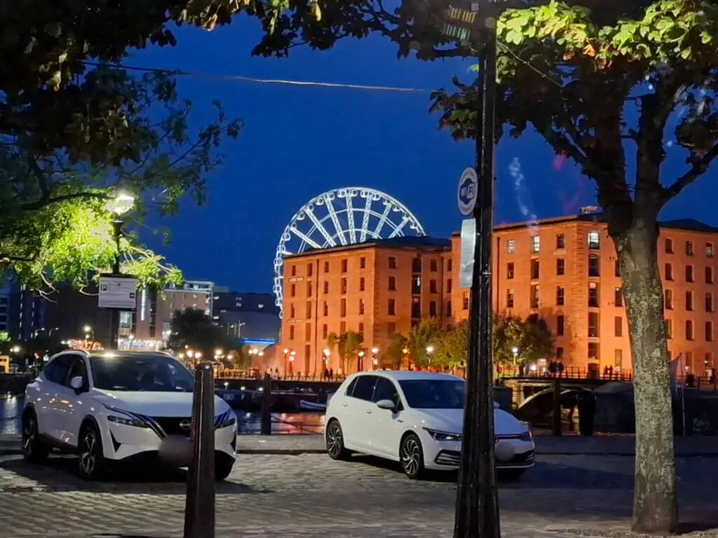 The Liverpool Eye ferris wheel lit up against a blue night sky and the square buildings of the Albert Dock warehouses which look orange in the fluorescent lighting