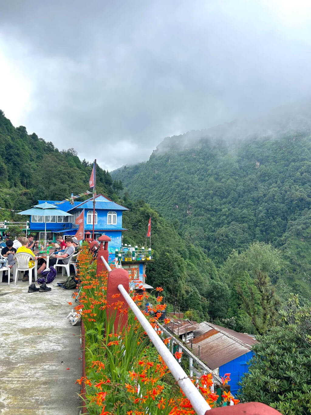 A bright blue painted building on the edge of a cliff, surrounded by green trees and clouds