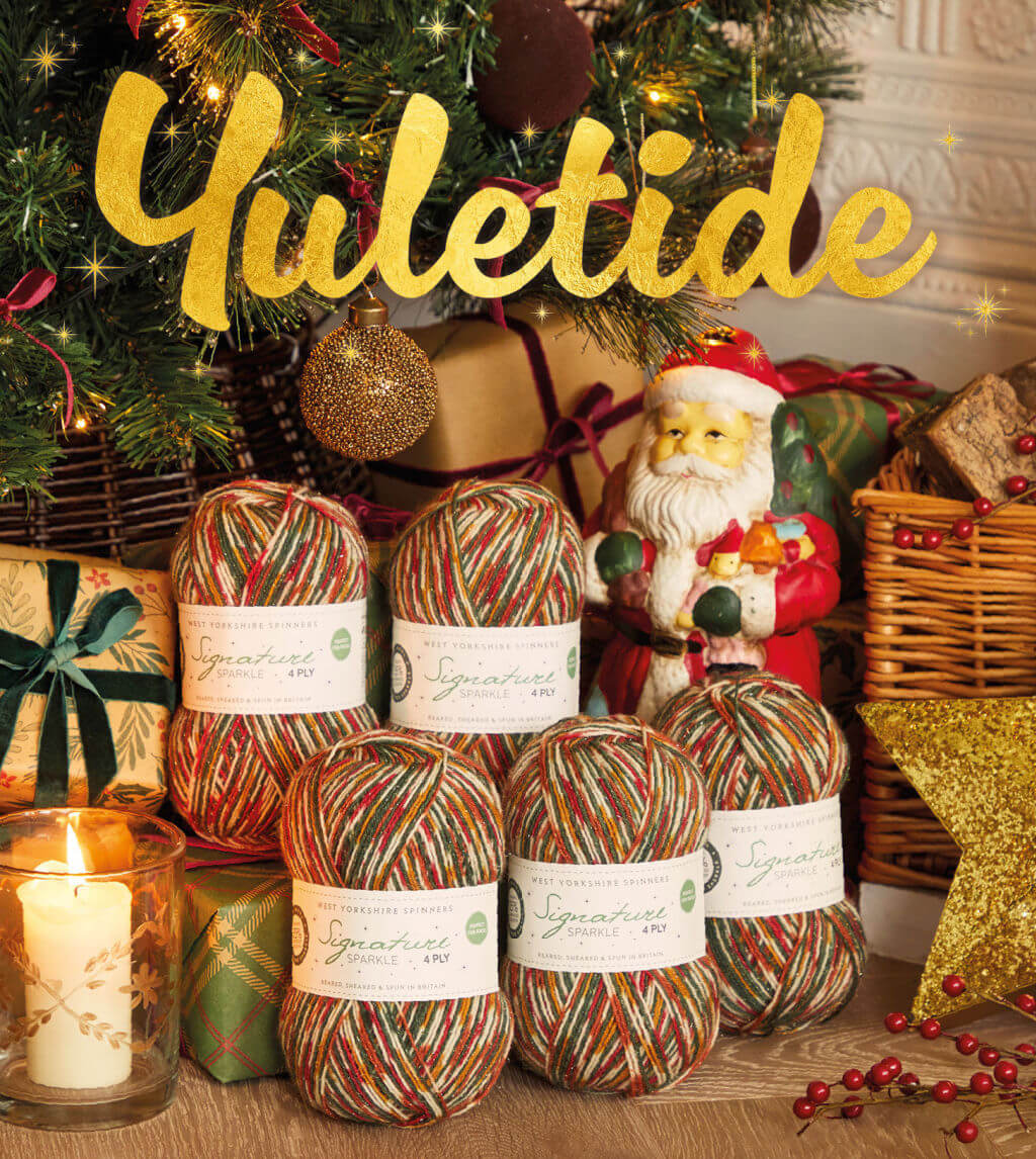 Five balls of yarn in shades of green, red and gold are stacked under a Christmas tree next to presents and a Santa ornament