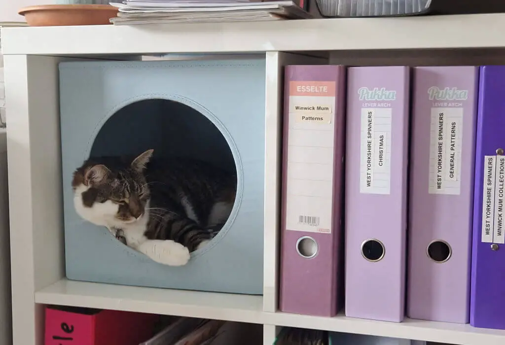 A tabby and white cat is sleeping in a blue box with a circular entry hole inside a square section of an IKEA Kallax unit