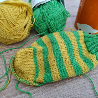 A partly-knitted green and yellow sock on a wooden table next to an orange mug of tea