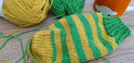 A partly-knitted green and yellow sock on a wooden table next to an orange mug of tea