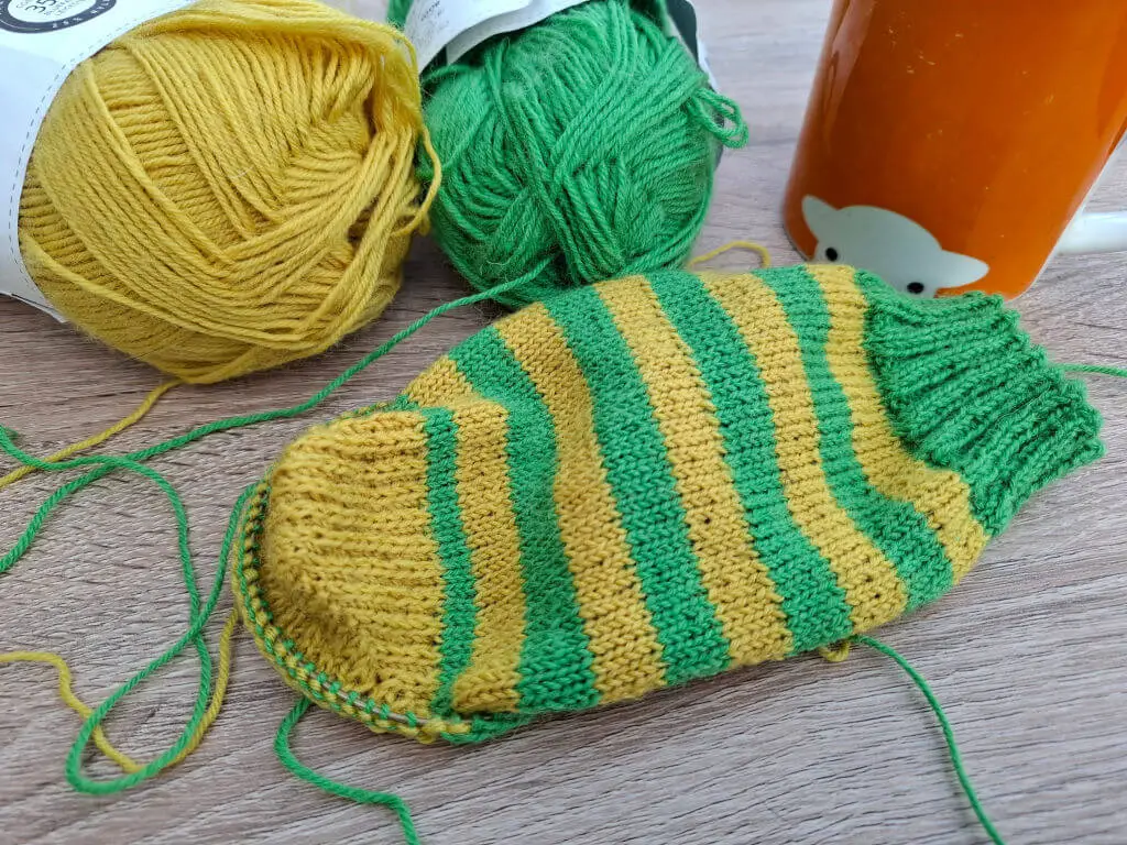 A partly-knitted green and yellow sock on a wooden table next to an orange mug of tea